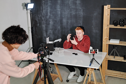 Young male vlogger in casual pullover and beanie showing his new photocamera during video shooting in studio
