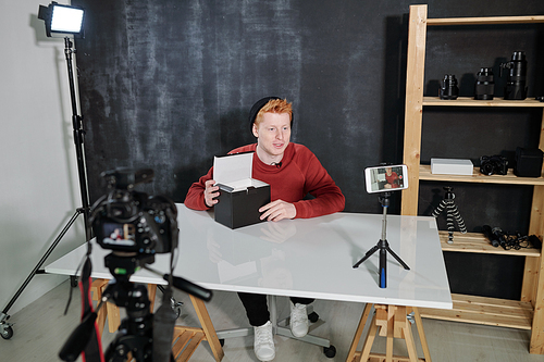 Young contemporary male vlogger going to unpack box with new photocamera during video shooting in studio