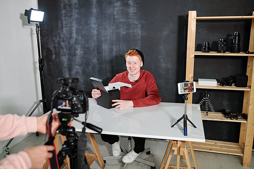 Cheerful male vlogger by desk going to unpack box with new photocamera during video shooting in studio