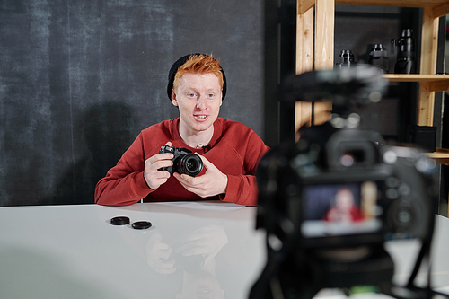 Cheerful young vlogger by desk holding new photocamera while talking about its characteristics during video shooting in studio