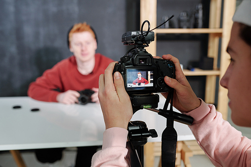 young video operator holding camera in front of male Vlog sitting by desk in studio and showing new photo equipment