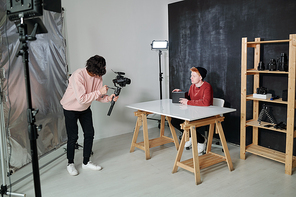 young video operator in casualwear holding camera in front of Vlog with black box sitting by desk in studio