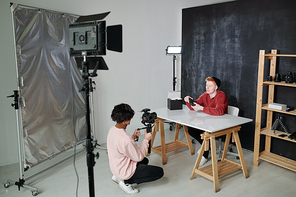 Young vlogger telling about new photo equipment while sitting by desk in front of operator with video camera