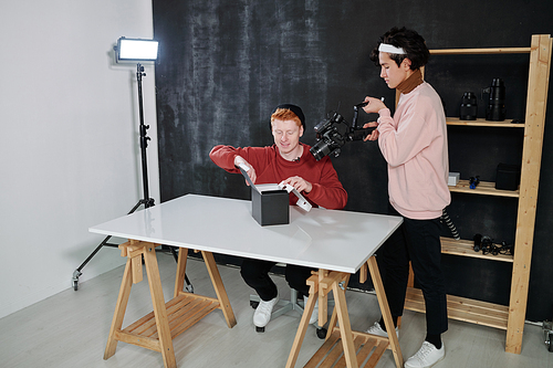 Young cameraman with video equipment shooting male vlogger sitting by desk and opening black box