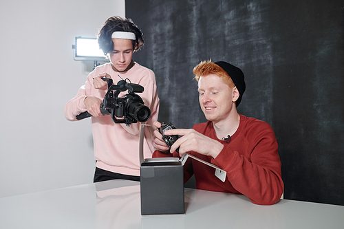 Young cameraman shooting happy male vlogger holding new photocamera over open box while sitting by desk