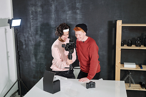 Two happy young male vloggers looking through new recorded video on digital screen of camera while discussing it in studio