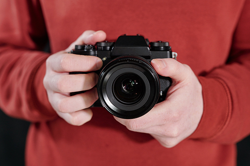 Hands of young male photographer in maroon sweatshirt holding new photocamera by his chest
