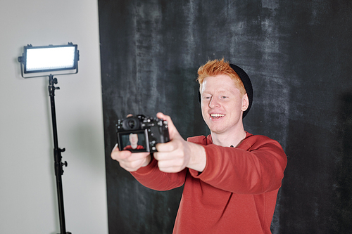 Cheerful young vlogger in casualwear standing against black background and holding camera in front of himself
