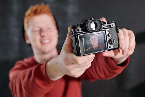 Hands of cheerful young vlogger holding camera in front of himself while posing in studio