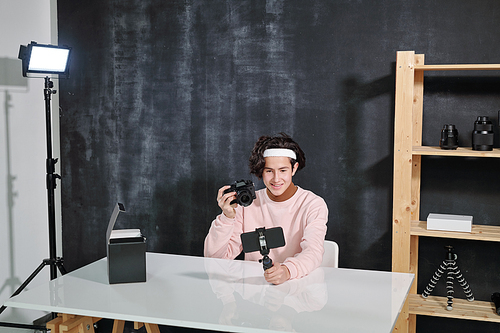 Happy guy with photocamera sitting by desk and shooting himself on smartphone which he holding in front