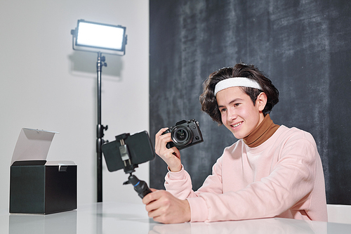 Young smiling male vlogger with photocamera sitting by desk and shooting himself on smartphone in studio