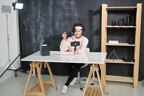 Young casual male vlogger sitting by desk and showing photocamera while shooting himself on smartphone in studio