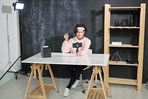 Young casual male vlogger sitting by desk and showing photocamera while shooting himself on smartphone in studio