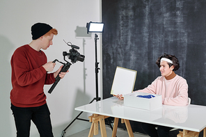 Serious young man with video shooting equipment standing in front of his friend sitting by desk and opening box with new footwear