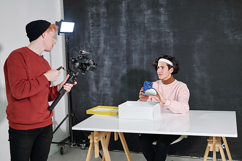 Young vlogger showing new footwear to his friend with video camera standing in front and shooting him in studio