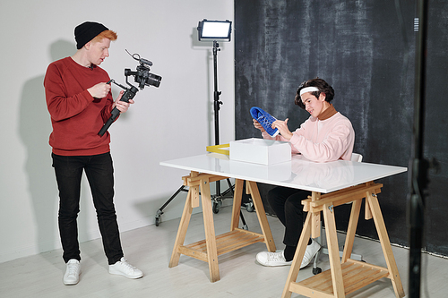 Young male vlogger looking at new footwear in front of his friend with video camera shooting him in studio