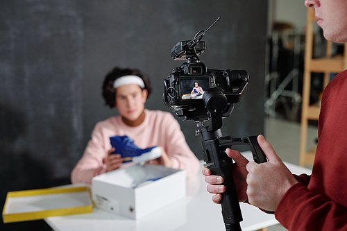 Young man holding video camera while standing in front of vlogger sitting by desk and telling about new footwear