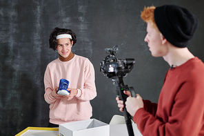 Smiling guy in casualwear holding new pair of sneakers over open box in front of camera held by his friend during shooting