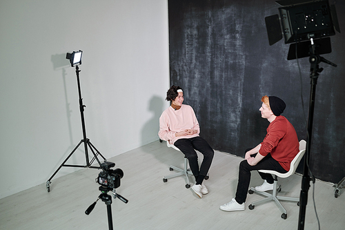 Two happy young vloggers in casualwear sitting on chairs in front of each other against black background in studio