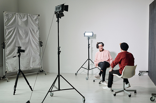 Two young male vloggers in casualwear sitting on chairs in front of each other in studio and interacting