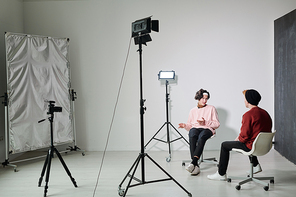 One of two young male vloggers explaining something to his friend while both sitting on chairs in front of each other in studio