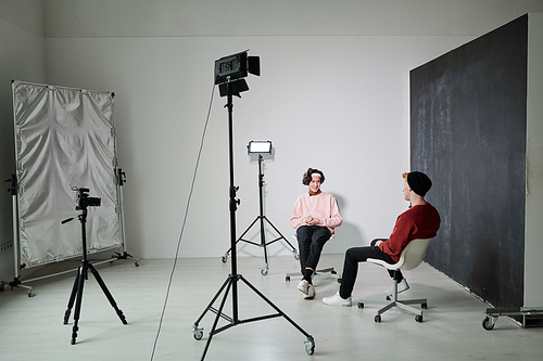 Two young friendly vloggers discussing contemporary fashion trends while sitting on chairs in front of each other in studio
