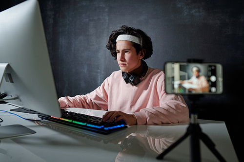 Young serious man in casualwear looking at computer screen while typing by desk in front of smartphone camera