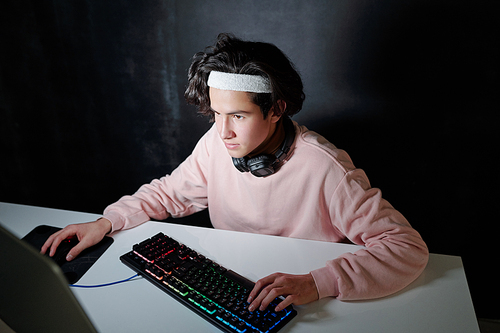 Young serious man in casualwear sitting by desk in front of computer screen while touching keys of keyboard