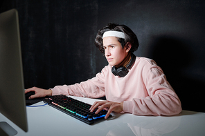 Serious guy with headphones around neck sitting in front of computer screen while concentrating on network in dark room