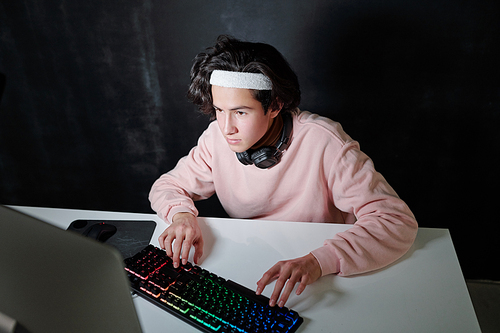 Serious teenager with headphones around neck pressing keys of keypad while sitting in front of computer screen in dark room