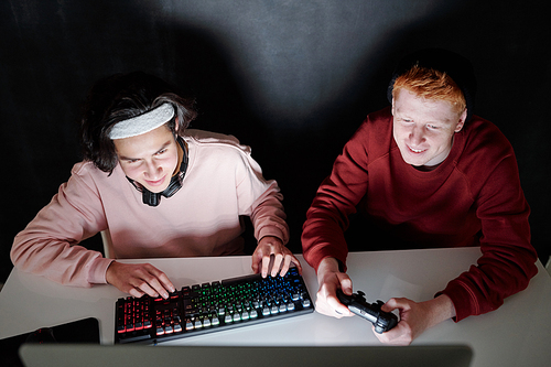 Two friendly teenagers in casualwear sitting by desk in front of computer screen while playing games in darkness