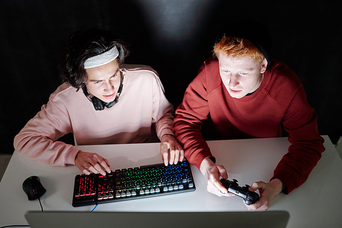 Two friendly guys in casualwear playing computer games while sitting by desk in front of monitor in darkness