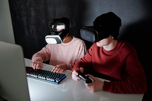 Two contemporary teenagers in vr headsets sitting by desk in front of computer screen while playing games in darkness