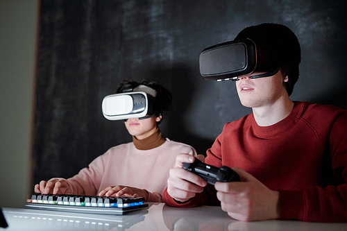 Two contemporary teenagers in vr goggles sitting in front of computer screen while playing games at night
