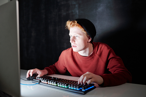 Contemporary teenager in sweatshirt and black beanie pressing keys of keypad and clicking mouse while sitting in front of computer screen