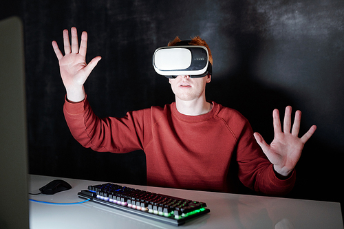 Casual young man in vr headsets sitting by desk in front of virtual computer screen in darkness