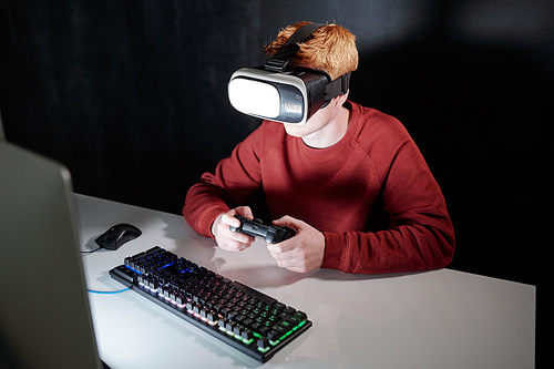 Young man with ginger hair in vr goggles sitting in front of computer screen while playing virtual games at night