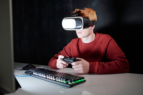 Young man with gamepad in vr headset sitting in front of computer screen while playing virtual games in darkness