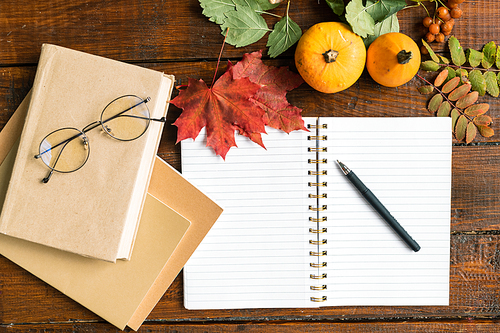 Overview of open copybook with blank pages and pen, stack of books with eyeglasses on top, two pumpkins and autumn leaves