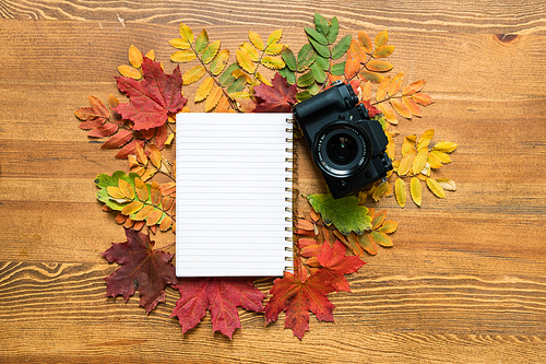 Wooden table with blank sheet of copybook surrounded by colorful autumn leaves with photocamera near by