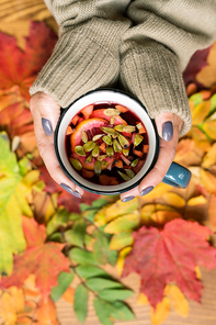 Female hands holding mug of hot herbal tea with lemon over multi-color autumn leaves