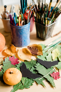 Composition of dry leaves, black paper bats, small pumpkin and two glasses with scissors and paintbrushes on table