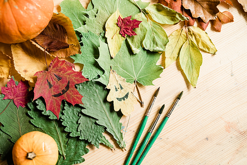 Composition of dry autumn foliage, gourds, halloween faces on leaves and paintbrushes on wooden table