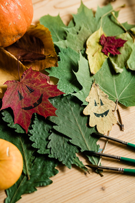 Overview of dry autumn foliage, drawn faces on leaves and three paintbrushes making halloween composition