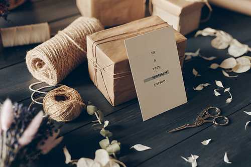 Close-up of card for special person leaning on packaged gift, scissors, twines, petals on wooden table