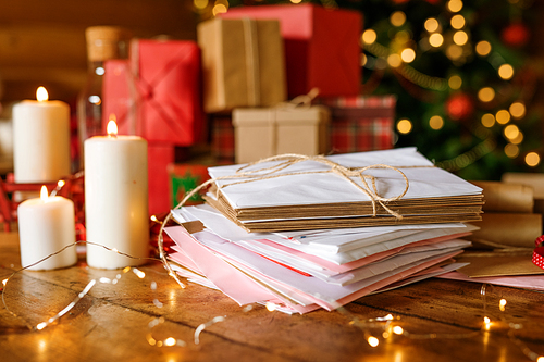 Stack of letters for Santa, three burning candles on wooden table and group of packed gifts on background