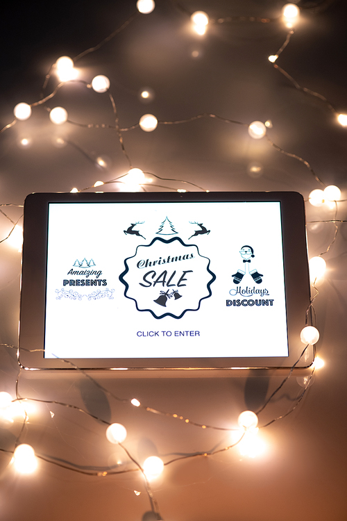 Digital tablet with online shop homepage on display surrounded by lit garlands that is a part of Christmas scene