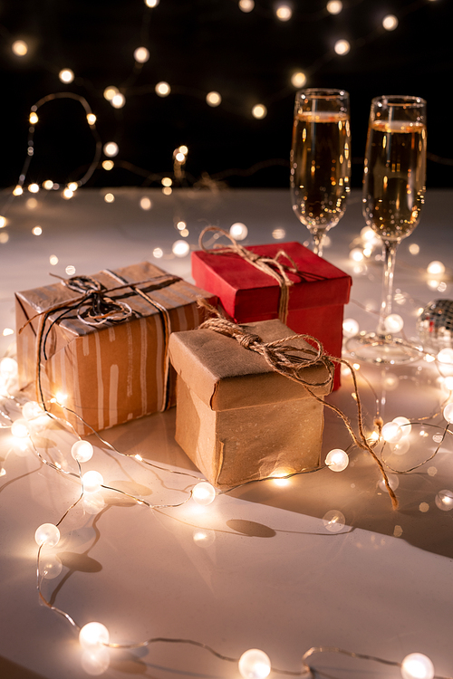 Three packed giftboxes, two flutes of champagne on table decorated with lit garlands for new year party