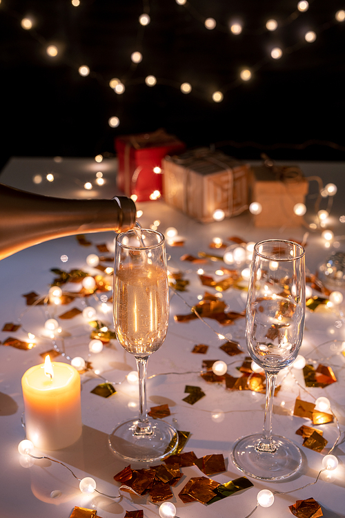 Two flutes and burning candle on table among golden confetti and lit garlands prepared for Christmas celebration