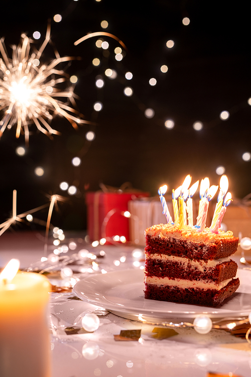 Piece of cake with many burning candles on plate surrounded by sparkling bengal lights, lit garlands and giftboxes